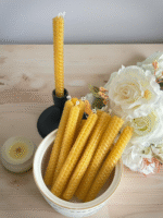 A set of hand-rolled beeswax taper candles displayed on a wooden surface, with one candle in a holder and the rest arranged in a ceramic bowl next to white artificial flowers—showcasing their natural honeycomb texture and warm golden color.